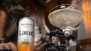 Lata de LAUÉ frizzante fría con gotas de condensación junto a una copa con bebida espumosa, sobre una mesa, mientras una persona juega rompecabezas al fondo en un ambiente íntimo y relajado.